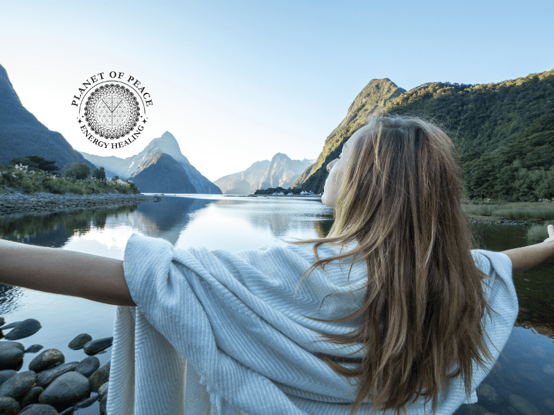 Woman standing peacefully by a river with arms open, symbolizing the emotional release of stubbornness and the flow of healing energy.